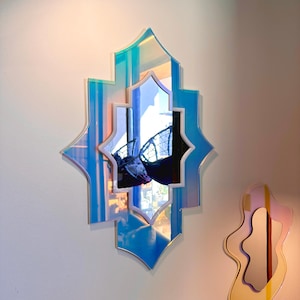 K&ouml;nnte beinhalten: Dekorativer Wandspiegel mit mehrschichtigem Design. Der Spiegel hat eine blaue und irisierende Oberfl&auml;che mit sternf&ouml;rmiger Form. Er reflektiert die Umgebung und erzeugt einen einzigartigen visuellen Effekt.