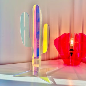 May include: A holographic iridescent cactus sculpture with a star-shaped base. The cactus is made of clear acrylic and has a pink and blue iridescent finish. The cactus is sitting on a white surface.