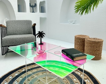 May include: A clear acrylic coffee table with a pink and green iridescent finish. The table is sitting on a woven rug with a black and white pattern. A book titled "sensizik" is on the table.