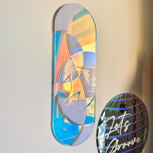 May include: A large, oval-shaped mirror with a geometric design in shades of blue, purple, and orange. The mirror has an iridescent finish and reflects the surrounding environment.