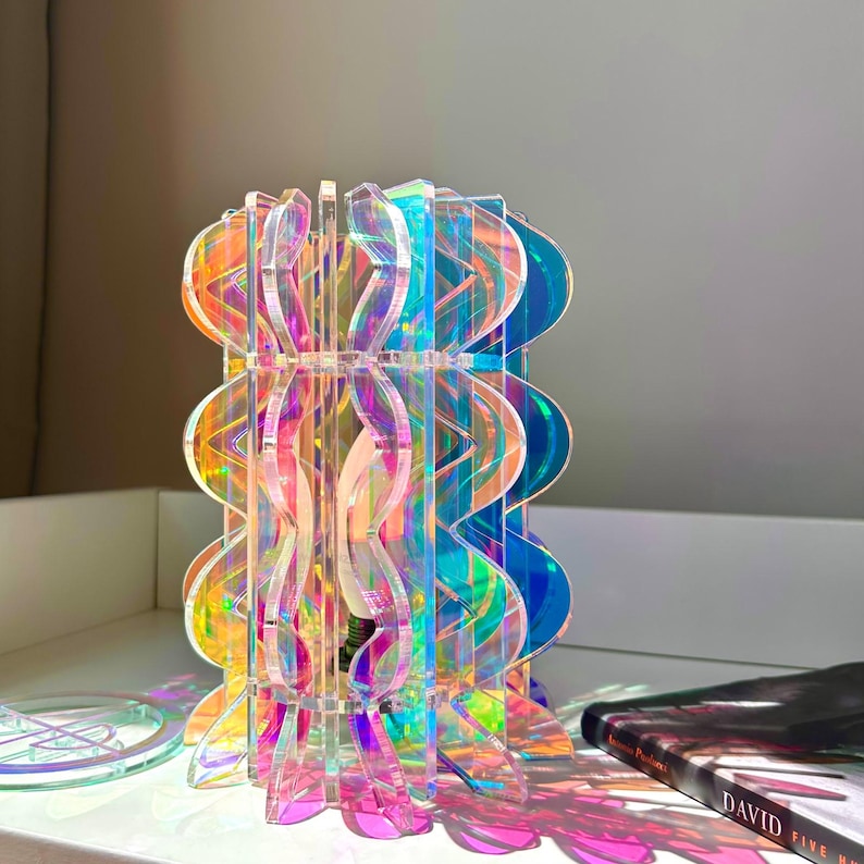 May include: A table lamp constructed from interlocking iridescent acrylic panels with a wavy, scalloped edge design. The translucent material reflects a spectrum of colors including pink, orange, and blue, creating a vibrant light display.