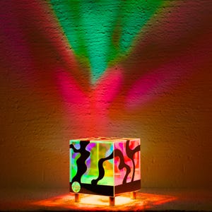 Könnte beinhalten: Eine schwarze, würfelförmige Lampe mit einem geometrischen Muster auf der Vorderseite. Die Lampe wirft ein farbiges Licht auf die Wand hinter ihr und erzeugt einen Regenbogeneffekt.
