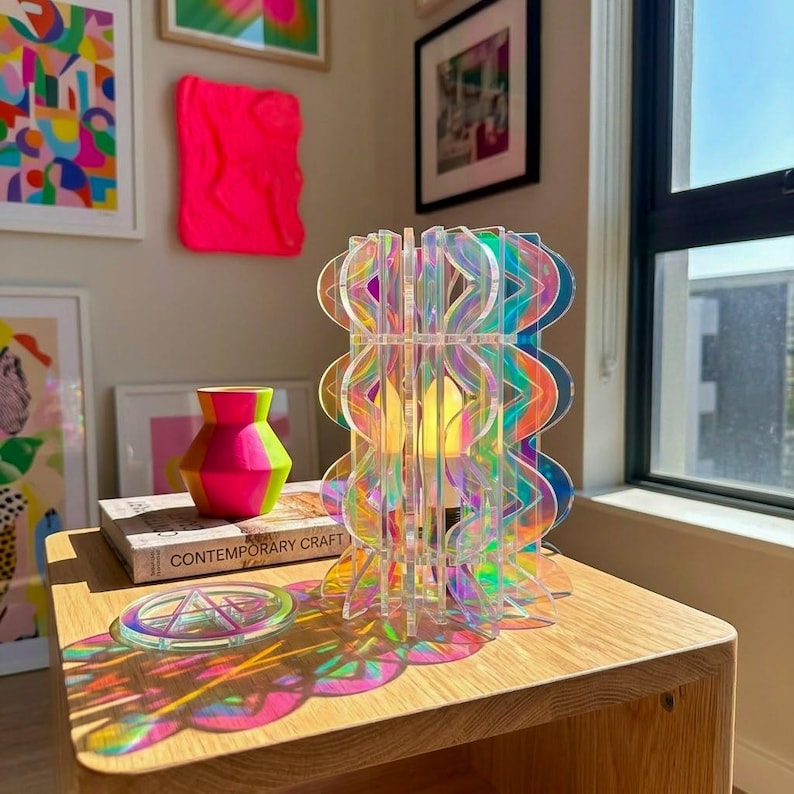 May include: A cylindrical lamp constructed from interlocking iridescent acrylic panels with a wavy, scalloped edge design. The lamp emits a warm glow from an internal light source, casting colorful, prismatic light reflections onto the wooden surface.