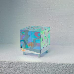May include: A small, iridescent cube-shaped table with a geometric pattern. The table has a mirrored finish and sits on four silver legs.
