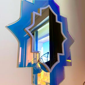 K&ouml;nnte beinhalten: Ein dekorativer Wandspiegel in schillerndem Blau und Gold. Der Spiegel hat eine einzigartige sternartige Form mit wei&szlig;em Rand und reflektierender Oberfl&auml;che. Der Spiegel ist an einer wei&szlig;en Wand befestigt.