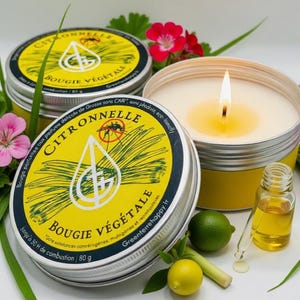 Könnte beinhalten: Eine brennende Citronella-Kerze in einer gelb-silbernen Dose, umgeben von rosa Blüten, grünen Blättern und einer kleinen Ölflasche. Zwei weitere Dosen mit "Citronnelle Bougie Végétale" sind ebenfalls zu sehen.
