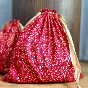 May include: Two red drawstring gift bags with a gold and white pattern of stars, hearts, and Christmas trees. The bags have beige drawstrings and are made of a soft fabric. The bags are perfect for holiday gifting.