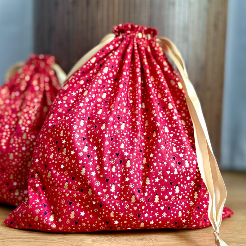 May include: Two red drawstring gift bags with a gold and white pattern of stars, hearts, and Christmas trees. The bags have beige drawstrings and are made of a soft fabric. The bags are perfect for holiday gifting.