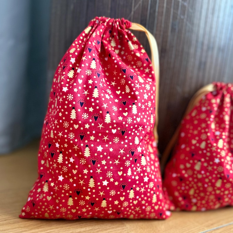 May include: Red drawstring gift bags with a festive pattern of gold stars, Christmas trees, and hearts. The bags have a cream-colored drawstring closure. The fabric has a repeating pattern of white dots.