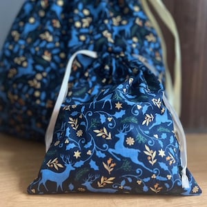 May include: Two blue drawstring bags with a white and gold floral pattern featuring stylized reindeer and holly. The bags are made of fabric and have a drawstring closure.