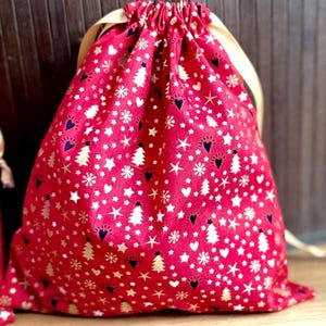 May include: A red drawstring gift bag with a festive pattern of white stars, hearts, snowflakes, and Christmas trees. Gold accents and a cream-colored drawstring complete the design. The bag is made of fabric.