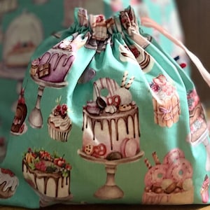 Può includere: Una borsa a cordoncino turchese con un motivo di torte e pasticcini colorati. La borsa è in tessuto e ha un cordoncino rosa.