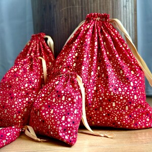 May include: Red drawstring bags with a gold and white holiday pattern of stars, trees, and hearts. The bags have a cream-colored drawstring closure. The bags are in various sizes, perfect for gift-giving or storage.