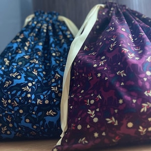 May include: Two fabric gift bags with drawstring closures. The bag on the left is blue with a gold deer and floral pattern. The bag on the right is purple with a gold deer and floral pattern.