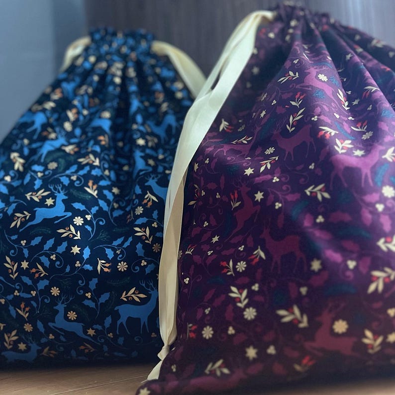 May include: Two fabric gift bags with drawstring closures. The bag on the left is blue with a gold deer and floral pattern. The bag on the right is purple with a gold deer and floral pattern.