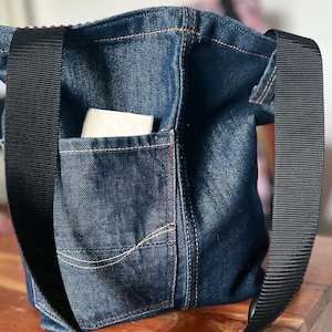 Puede incluir: Una bolsa de lona de mezclilla azul oscuro con correas negras y un bolsillo grande en la parte delantera. La bolsa tiene un pañuelo blanco dentro del bolsillo.