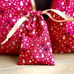 May include: Red drawstring gift bags with a festive pattern of gold and white stars, hearts, snowflakes, and Christmas trees. The bags have a cream-colored ribbon closure, perfect for holiday gifting.