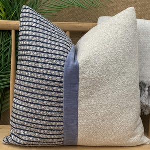 Könnte beinhalten: Zwei dekorative Zierkissen. Ein Kissen hat einen strukturierten, hellgrauen Stoff. Das andere Kissen hat auf einer Seite ein blau-weißes Streifenmuster und ein durchgehendes blaues Feld. Die Kissen liegen auf einer Holzbank.