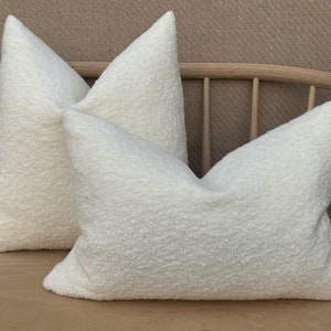 May include: Two white fluffy throw pillows with a textured surface. The pillows are arranged on a wooden bench.