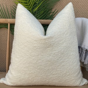 May include: A white, fluffy pillow with a textured surface. The pillow is sitting on a wooden bench with a gray throw blanket with fringe.