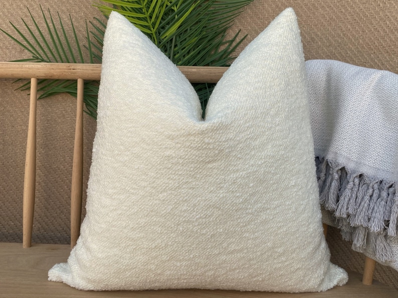 May include: A white, fluffy pillow with a textured surface. The pillow is sitting on a wooden bench with a gray throw blanket with fringe.