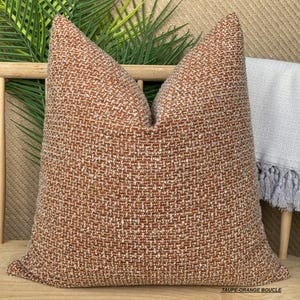 May include: A brown and white patterned throw pillow with a textured fabric. The pillow is on a wooden chair with a white throw blanket with fringe.