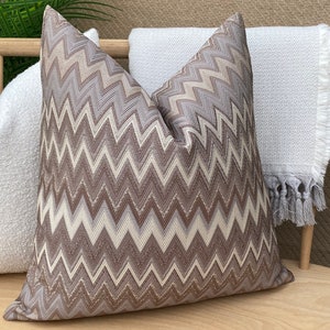 Puede incluir: Una almohada decorativa con un patrón de chevron marrón, blanco y beige. La almohada está hecha de una tela suave y texturizada.