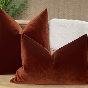 Puede incluir: Tres almohadas de terciopelo de color óxido dispuestas en un banco de madera. Las almohadas son todas del mismo tamaño y forma, y tienen un tacto suave y lujoso.