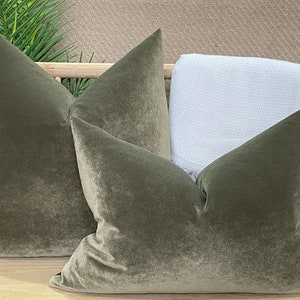 May include: Two green velvet pillow covers with a subtle sheen. The pillows are arranged on a wooden surface with a white throw blanket behind them.