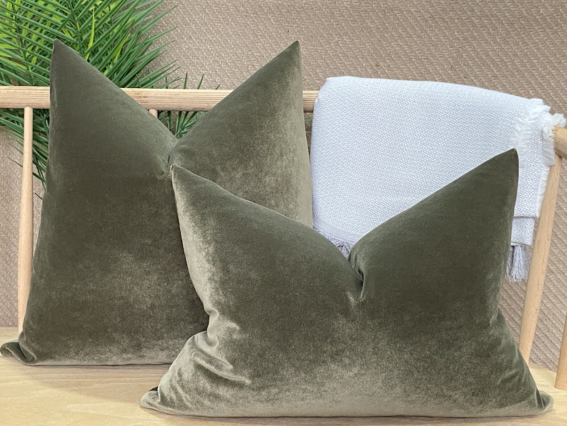 May include: Two green velvet pillow covers with a subtle sheen. The pillows are arranged on a wooden surface with a white throw blanket behind them.