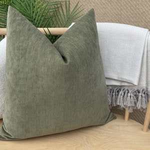 May include: A green velvet throw pillow with a subtle texture sits on a wooden chair with a white throw blanket with fringe.
