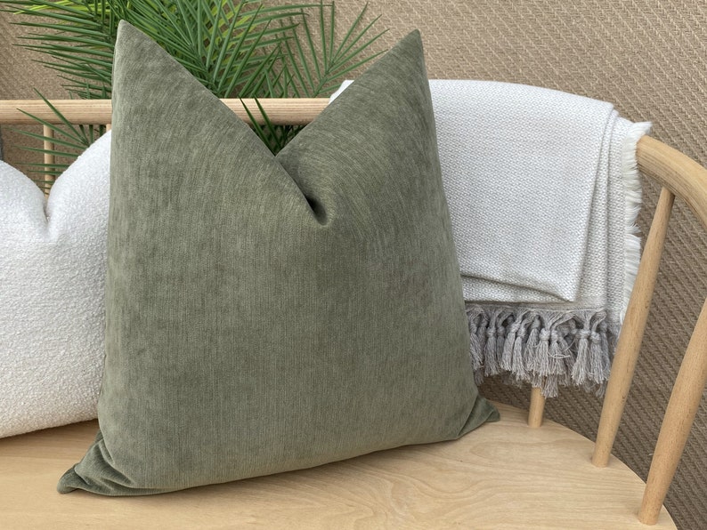 May include: A green velvet throw pillow with a subtle texture sits on a wooden chair with a white throw blanket with fringe.