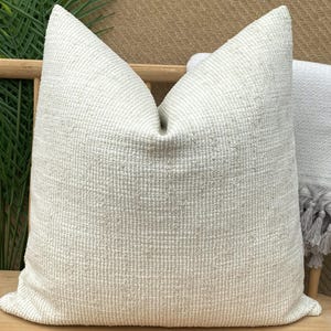 Op de afbeelding: Een groot, crèmekleurig, getextureerd kussen met een geweven uiterlijk. Het kussen is vierkant en heeft een zachte uitstraling. Het is geplaatst op een houten stoel met een grijze deken met franjes.