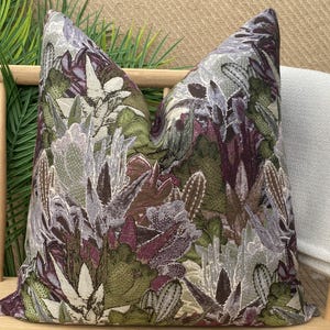 May include: A decorative pillow with a floral pattern featuring a variety of green, purple, and white flowers. The pillow is made of a textured fabric and has a square shape.
