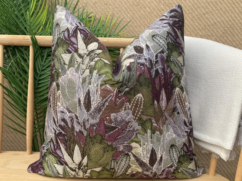 May include: A decorative pillow with a floral pattern featuring a variety of green, purple, and white flowers. The pillow is made of a textured fabric and has a square shape.