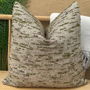 May include: A decorative square pillow with a textured, woven fabric in shades of gray, beige, and olive green. The pillow is placed on a wooden chair with a gray blanket draped over it. The pillow's design features a subtle, linear pattern.