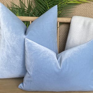 Könnte beinhalten: Zwei hellblaue Samtkissen mit weicher, strukturierter Optik. Die Kissen liegen auf einer Holzbank, mit einer hellen Decke und Grünzeug im Hintergrund. Die Kissen sind ein gedämpftes, pastellfarbenes Blau.