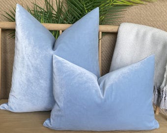 Könnte beinhalten: Zwei hellblaue Samtkissen mit weicher, strukturierter Optik. Die Kissen liegen auf einer Holzbank, mit einer hellen Decke und Grünzeug im Hintergrund. Die Kissen sind ein gedämpftes, pastellfarbenes Blau.