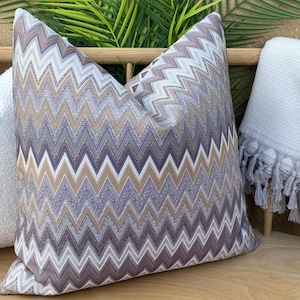 May include: A patterned cushion featuring a chevron design in purple, white, and tan hues. The cushion is displayed on a wooden surface alongside other cushions and a fringed blanket. The design adds a modern touch to home decor.