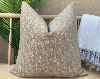 May include: A beige throw pillow with a subtle white textured pattern. The pillow is sitting on a wooden bench with a light blue throw blanket with tassels.