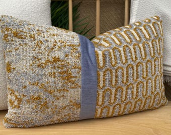 May include: Decorative rectangular pillow with a textured gray and gold pattern on one side and a geometric design on the other. A vertical strip of blue fabric divides the two sides. The pillow is a home decor item.