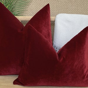 May include: Two red velvet throw pillows with a square shape and a subtle sheen. The pillows are arranged on a wooden surface.