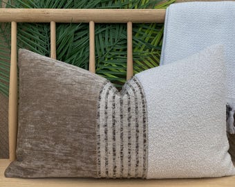 Puede incluir: Un cojín decorativo con un diseño texturizado. Presenta una tela marrón aterciopelada en un lado, una franja tejida en el centro y una tela gris claro texturizada en el otro lado. El cojín es rectangular y descansa sobre una superficie de madera.
