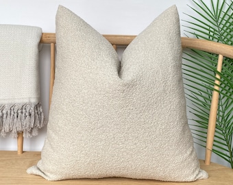 May include: A large, textured, light beige pillow with a soft, nubby surface. The pillow is placed on a wooden bench next to a fringed throw blanket. The pillow is a neutral color, suitable for various home decor styles.