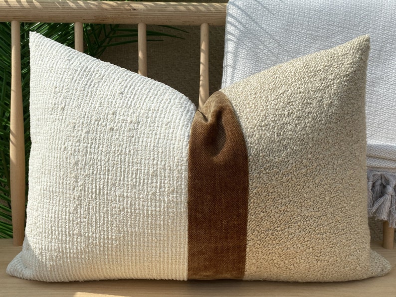 Puede incluir: Una almohada de tiro blanca y beige con una franja de terciopelo marr&oacute;n en el centro. La almohada est&aacute; hecha de una tela texturizada y tiene un aspecto ligeramente desgastado.