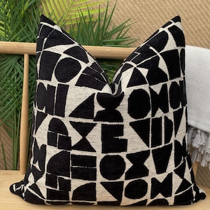 May include: A cushion with a black and white geometric design. The pattern includes various shapes like circles and triangles. The cushion is on a wooden bench. The cushion's dimensions are approximately 40cm x 40cm.