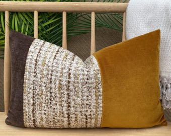 Puede incluir: Un cojín decorativo rectangular con un diseño de bloques de color. El cojín presenta un extremo de terciopelo marrón oscuro, un centro texturizado con hilos beige, marrón y dorados, y un extremo de terciopelo amarillo mostaza. El cojín mide unos 50 cm de largo.