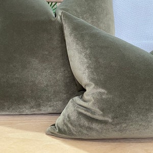 May include: Two green velvet cushion covers with a soft, plush texture. The cushions are arranged on a light wood surface.