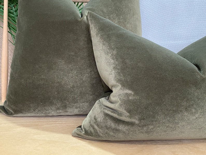 May include: Two green velvet cushion covers with a soft, plush texture. The cushions are arranged on a light wood surface.