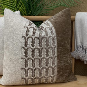 May include: A selection of decorative cushions in neutral colours. One has a textured off-white fabric, another a geometric pattern in brown and beige, and the third is a brown velvet.